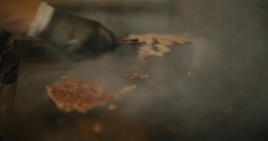 Man flips ground meat patties on smoking griddle before topping them with cheese slices