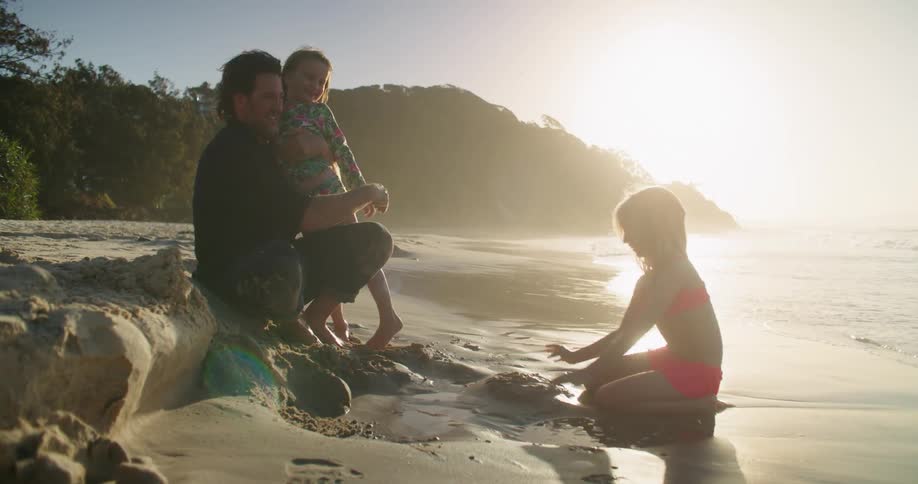 A man holds his toddler daughter on the beach as another daughter plays in the sand