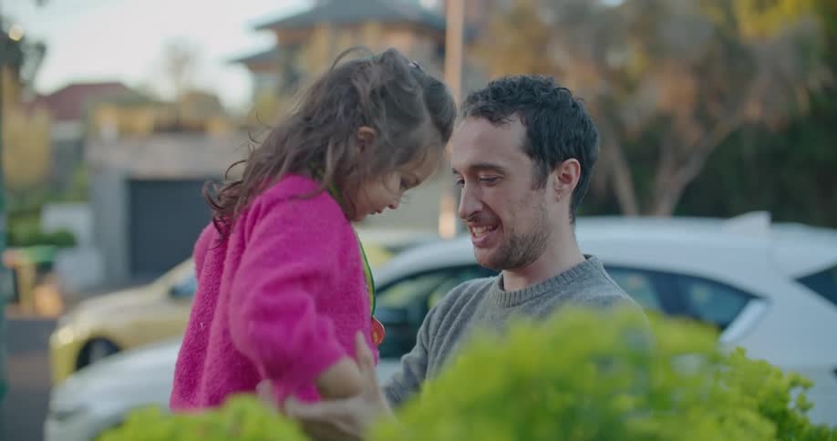 Father helps daughter balance near flower bed as traffic passes on road in background