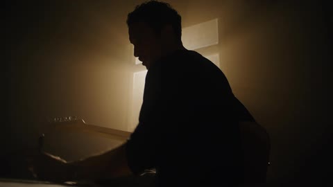 Man composes for guitar while sitting on bed in shadowy room partly silhouetted by window light