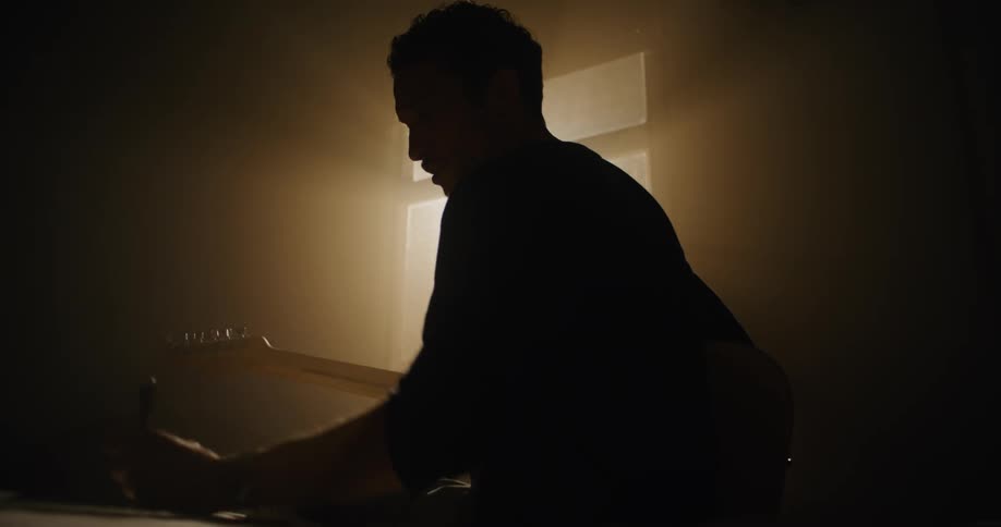 Man composes for guitar while sitting on bed in shadowy room partly silhouetted by window light
