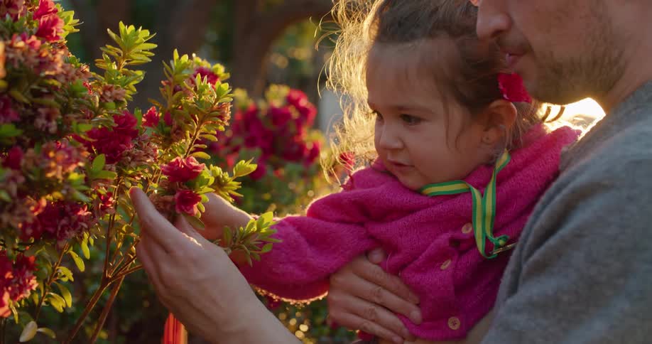 Man with grey sweater and earring holds toddler girl with pink coat picking up flower
