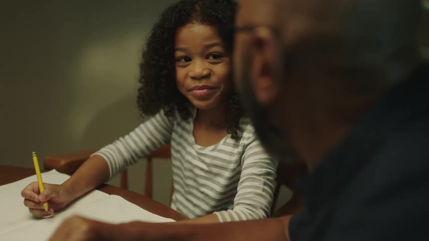 A young girl sits at a table doing homework with a man beside her