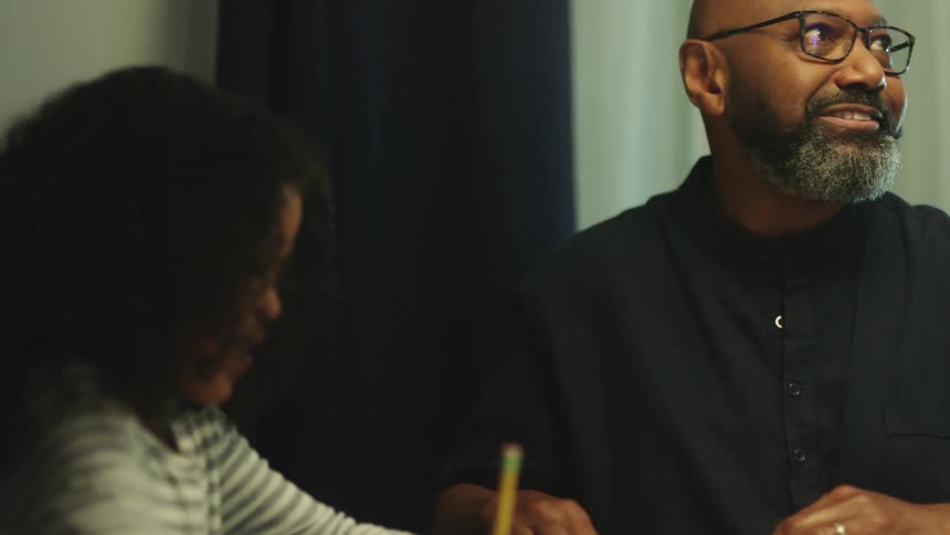 A father watches his daughter do her homework while indoors and sitting at a table