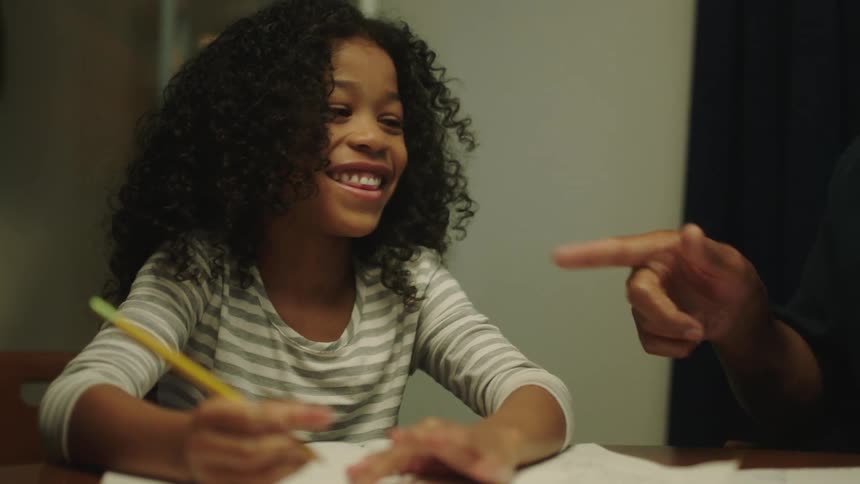 A young girl is sitting at a desk and writing in a notebook while laughing with a teacher