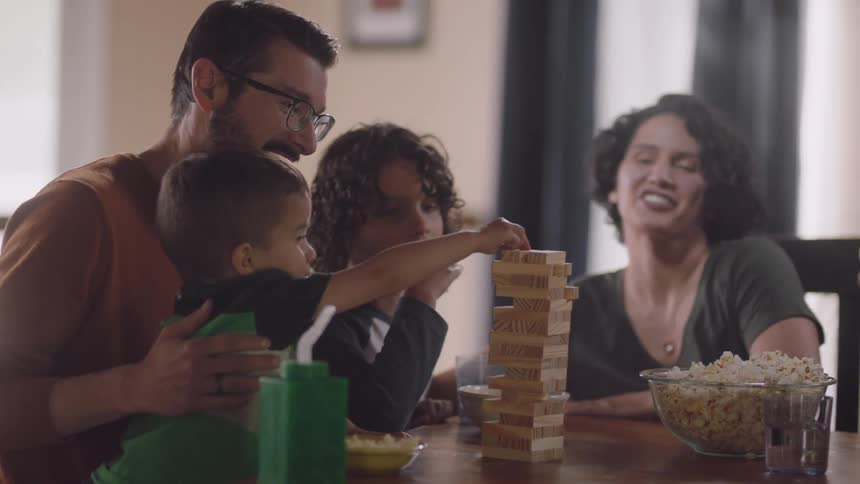 A family of four are sitting in their home at a table with popcorn and playing a game in the daytime