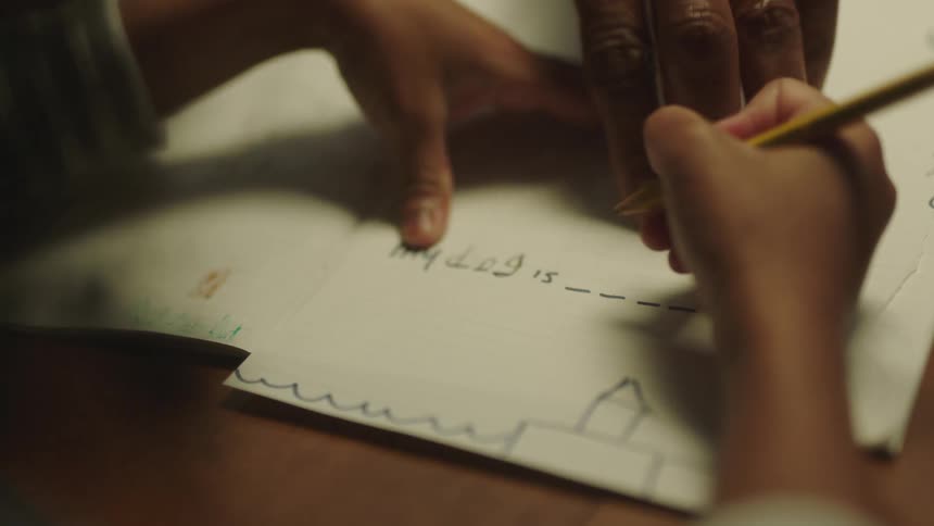 A child is holding a pencil writing on paper and an adult is pointing to writing on the paper