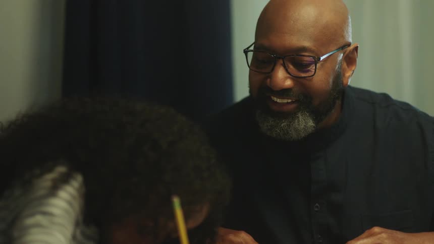 A dad smiles and nods as he watches his daughter do her homework at the table