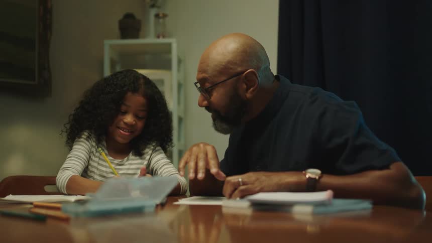 A father helps his daughter work on homework at a table covered in books