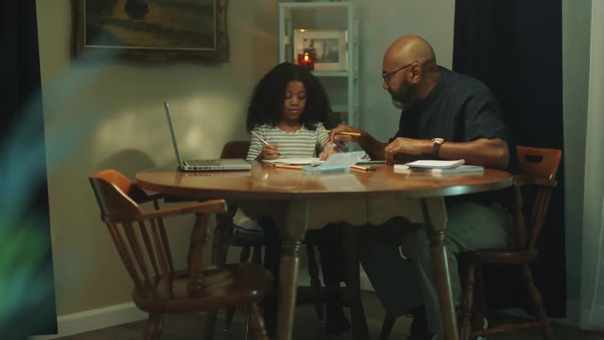 A father is helping his daughter with homework as they sit at a table