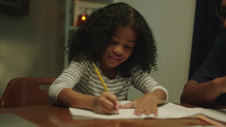 A young girl is sitting at a table writing with a pencil and laughing as a man next to her is watching