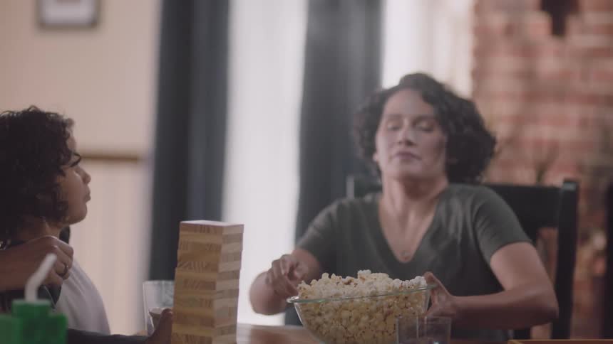 Mother with curly hair brings popcorn to father and sons playing jenga at table