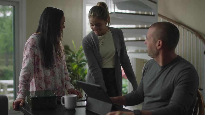 A teenager sets down her backpack and greets her parents as her father works on a laptop at home