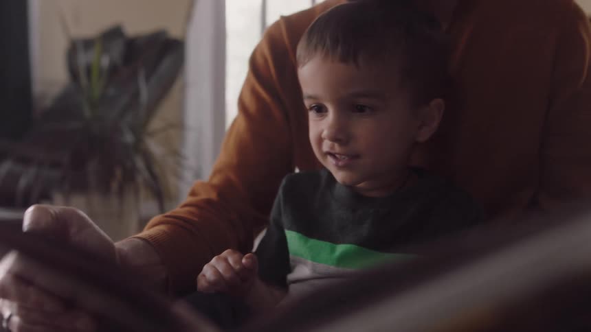 A little boy sits in his fathers lap at home as he reads a book to him
