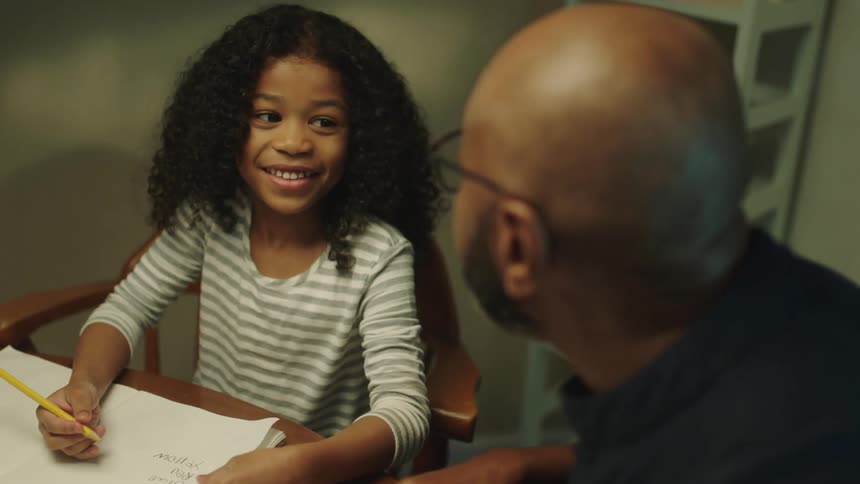 A father and daughter sit at a table as she does homework in a notebook