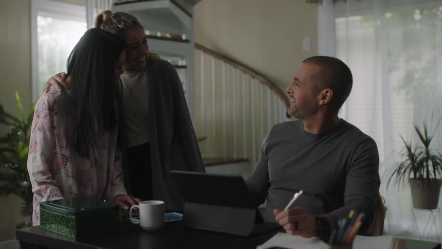 A man is sitting in his home at a desk talking to a woman as a teenage girl comes down the stairs and hugs her and leaves