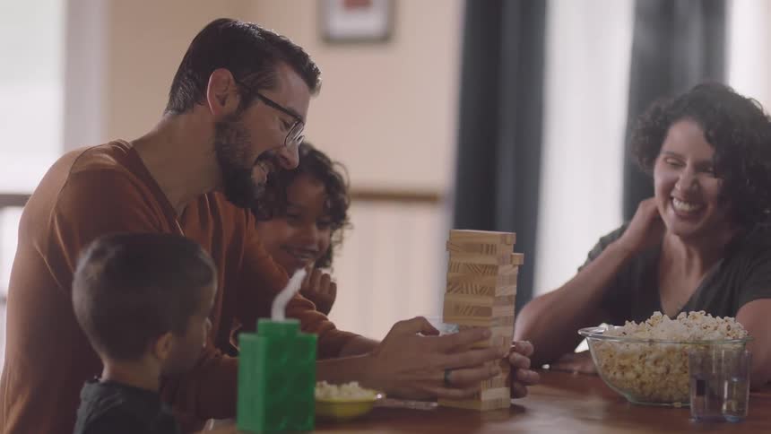 Dad taps at a jenga block while mom and the kids laugh and eat popcorn