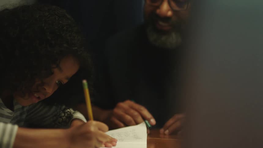 A young girl sits at a table doing homework with a man beside her