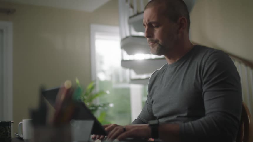 Man with short hair and gray sweatshirt sits at table and uses tablet smiling