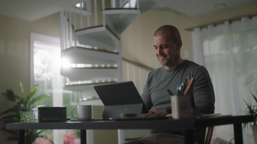 A man types on his laptop while sitting at home in front of a spiral staircase
