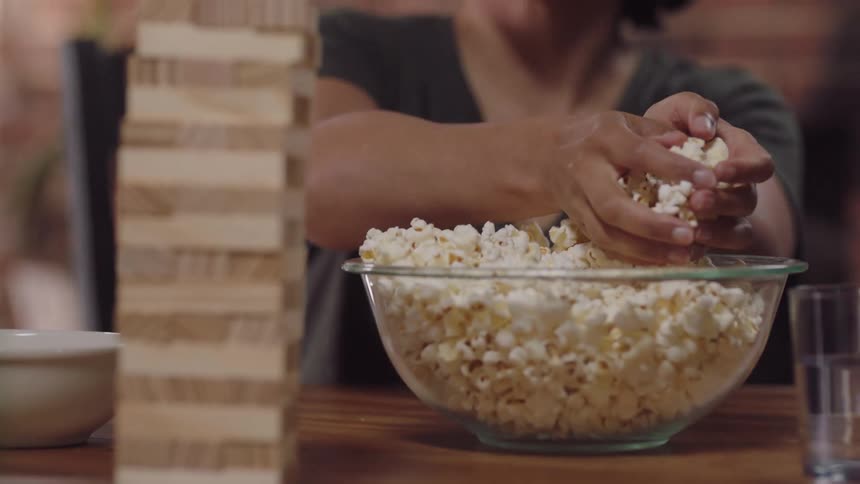 Woman with curly hair and grey t shirt puts popcorn in bowl next to cenga on the table