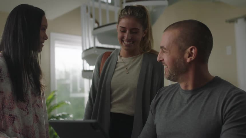 A family laugh together while gathered around the fathers desk as he types on a laptop