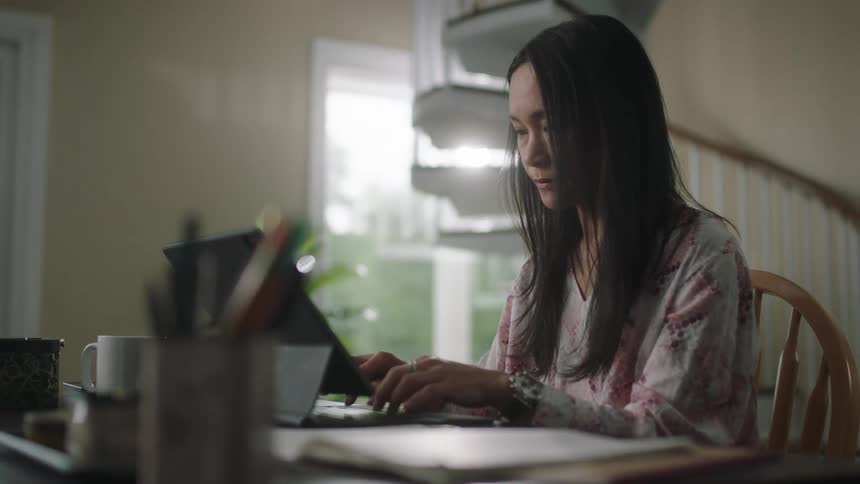 Woman sits at table near spiral staircase typing on computer