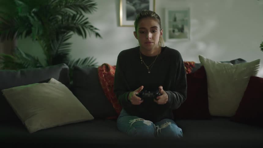 A woman is sitting on a couch in a home while holding a game controller and playing a video game during the day