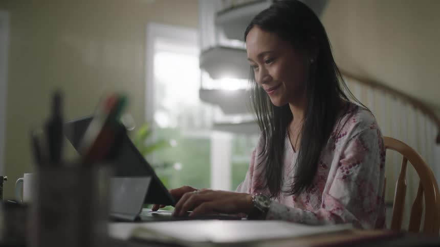 Woman with straight hair and pink white shirt sits at table and types on tablet keyboard in living room at home