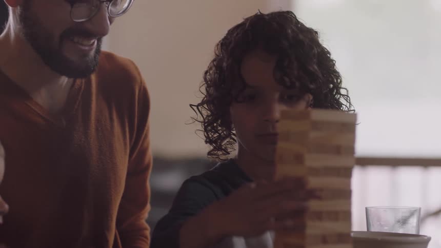 A young boy moves a piece on a jenga game as his family sits at the table and plays the game together while eating popcorn