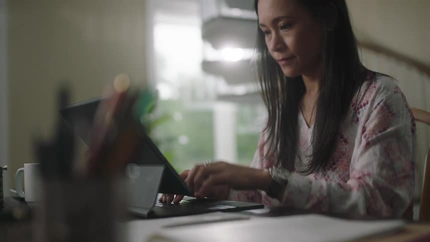 A woman sits at a table typing on a keyboard and touches the screen of a tablet