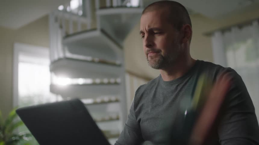 A man sits at home and thinks and then laughs as he uses his laptop while a woman sits at the table with him
