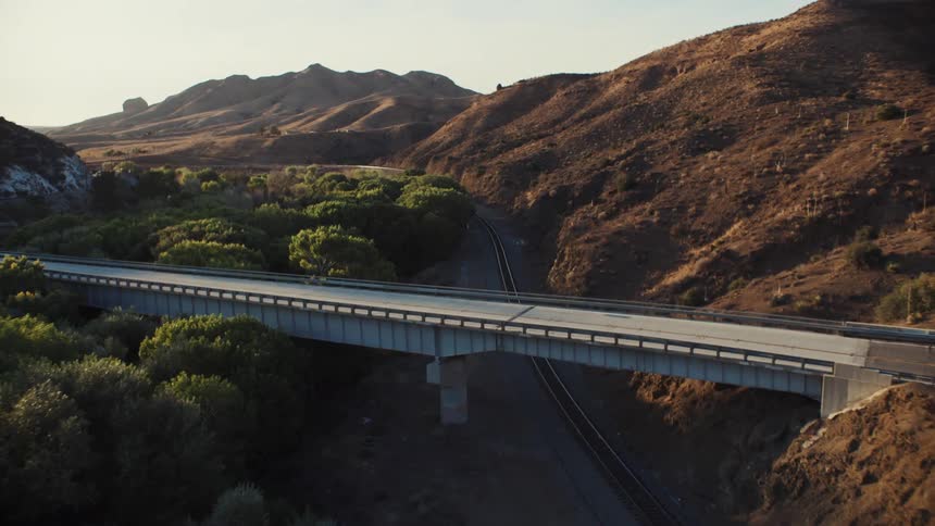 Road crosses along bridge over train tracks between forest and hills