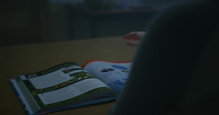 A young woman is sitting at a table with an open book in a classroom during the day