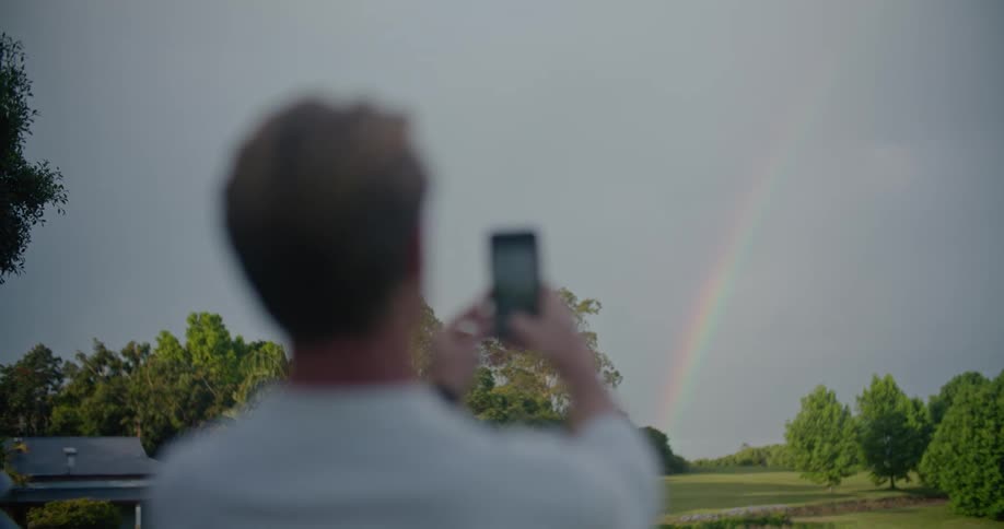 A man is outside taking pictures of a rainbow with his phone