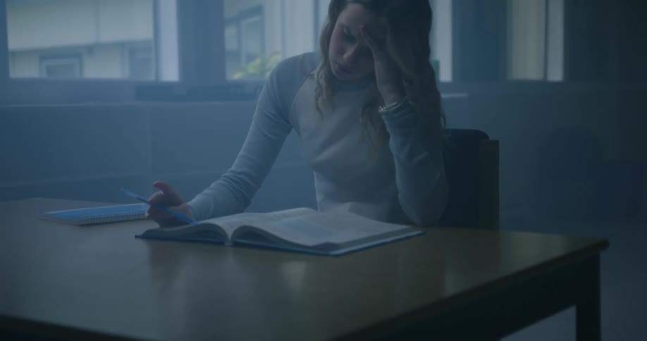 Young woman sitting at a desk in a dimly lit room reads a book while studying