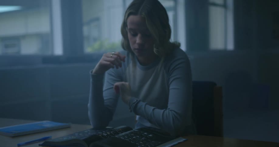 A woman rubs her head as she sits at a table looking at a book