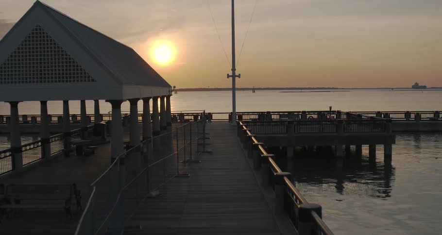 The sun glows golden in the sky above the water near an empty pier with tables and chairs