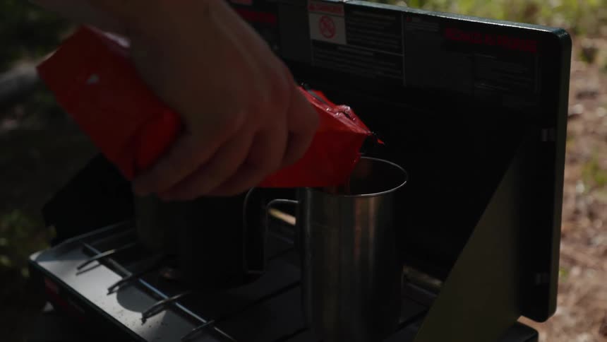 A man is pouring coffee grinds into a cup on an outdoor grill in a backyard at night