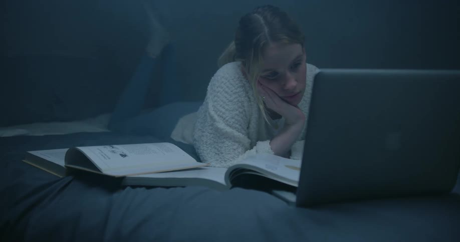 A girl is lying on her bed with a laptop computer and two open books