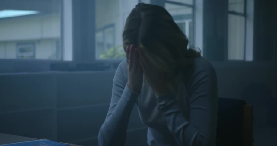 Young tired girl sitting inside a dark and dimly lit room reading a book rubs her forehead with her fingers