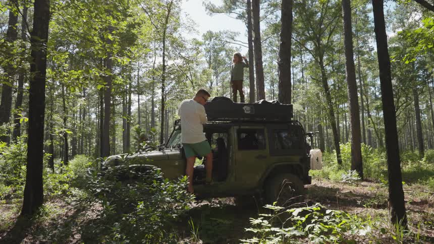 A dad secures supplies under a tarp on the car while his child walks on top of the jeep