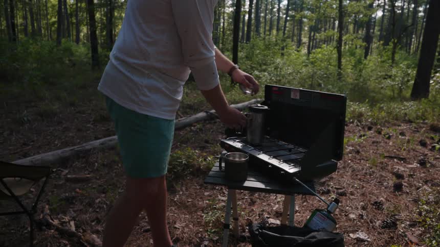 A man pours liquid from a teapot into a mug at a campsite grill