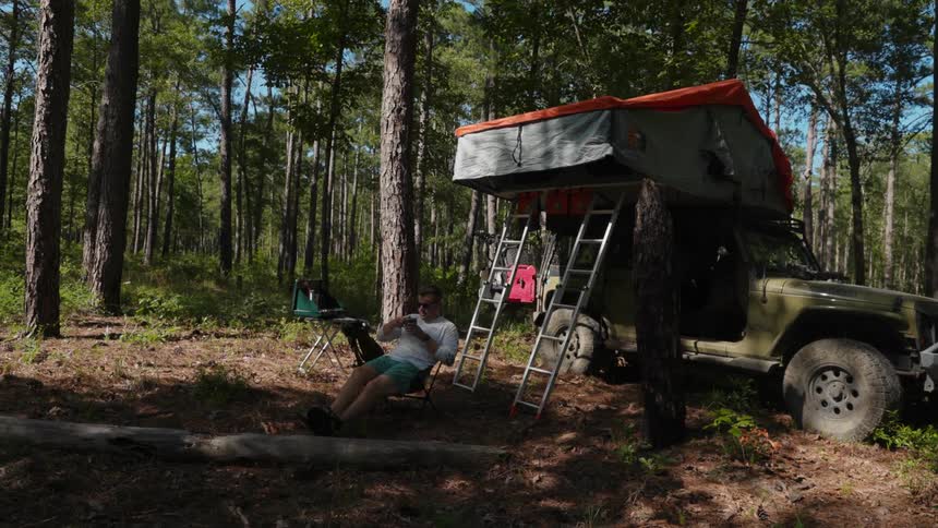 Man camping in the forest sits down on a foldable chair near a jeep with a tent on the roof