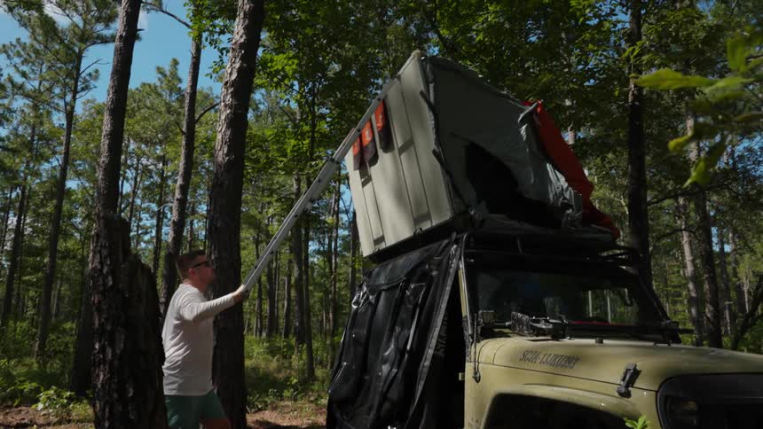 Man unfolds tent built onto roof of jeep parked in forest
