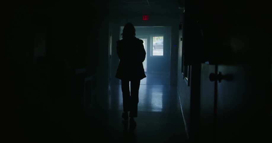 A woman walks down a dark hallway towards a glass door