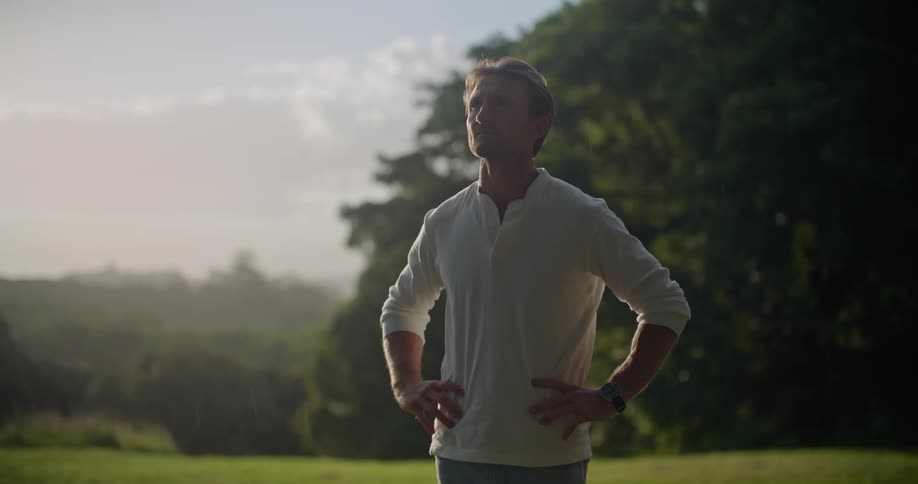 A man is standing in a grass field in a woodland area and realizes that its starting to rain