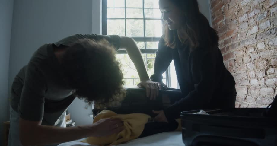 A man stuff clothing into a suitcase while a friend helps him get it closed