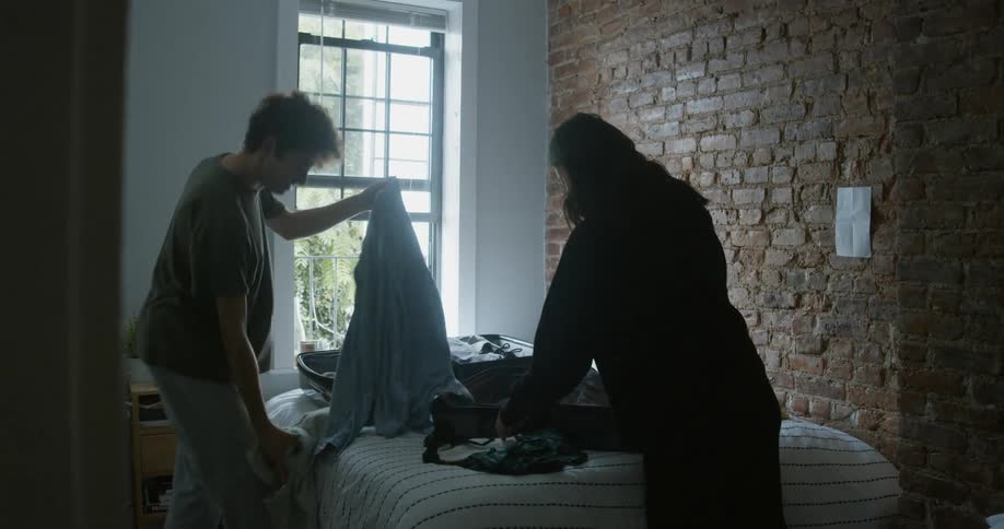 A man and woman fold up clothes while packing in their bedroom