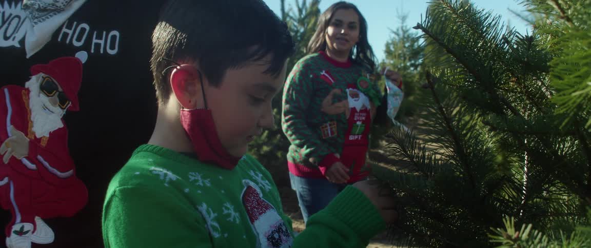 Family wearing christmas sweaters and face masks walk down aisle of tree farm